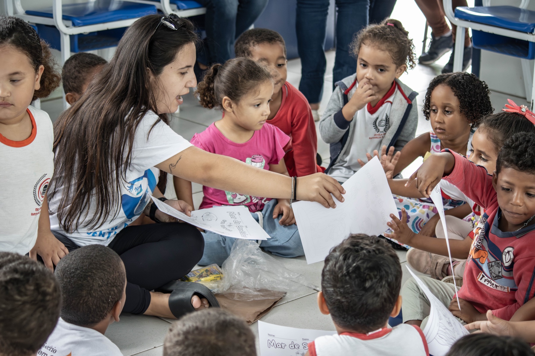 Educadora distribui atividades para um grande grupo de crianças sentado em roda