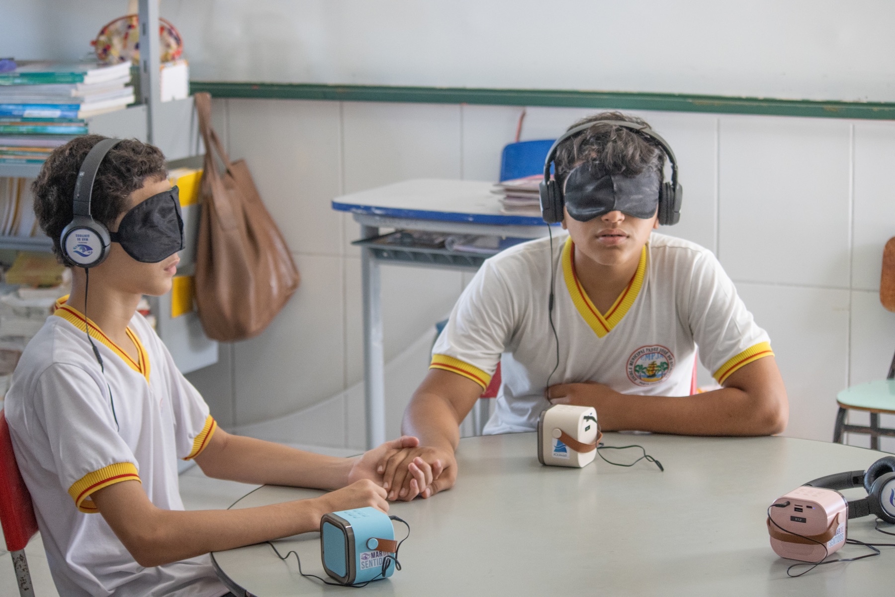 Dois estudantes vendados usam fones de ouvido e seguram as mãos durante experiência sensorial